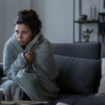 Person wrapped in a blanket sitting on a sofa because one room in their home feels colder than the rest during winter.
